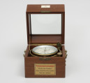 Chronometr na zawieszeniu Cardana - ujęcie z przodu; Chronometr w drewnianej skrzynce z kluczem. Skrzynka drewniana z mahoniu, lakierowana. Wieczko skrzynki z szybą fazowaną; boki podstawy skrzynki zaopatrzone w mosiężne ucha. Od frontu zamknięcie typu zapinka w formie okucia, na wieku płaskie, u podstawy zaopatrzone w trzpień. Powyżej okucia biała, plastikowa tabliczka znamionowa o wymiarach 4,3x2,7 cm, poniżej nalepka informacyjna. Z lewej strony dolnej części skrzynki mosiężna, dokręcana śrubami prowadnica, zabezpieczającą wieko przed zamknięciem i wyłamaniem. Krawędzie zamknięcia skrzynki z frezowaniem typu pióro-wpust, służące uszczelnieniu. 
Cyferblat chronometru biały z czarnymi oznaczeniami. Godziny w rzymskim zapisie liczbowym, obwiedzione podziałka minutową. Cyfry tarczy sekundowej i czasu pracy arabskie. Wskazówki, godzinowa w formie strzałki i prosta minutowa, wykonane z mosiądzu. Wskazówka sekundnikowa i czasu pracy z pomalowanego na czarno metalu. Podziałka wskaźnika godzin czasu pracy w formie niepełnego okręgu, umieszczony pod rzymską godziną XII. Mosiężny klucz o okrągłym uchwycie na dłoń w górnej części posiada otwór.