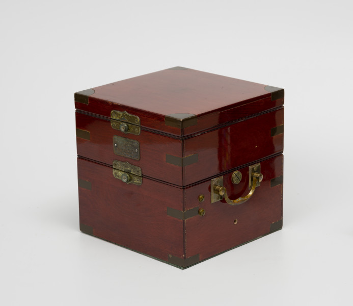 chronometr morski Gost - Ujęcie z przodu; Skrzynka drewniana kwadratowa w kolorze ciemnej wiśni, lakierowana na wysoki połysk. Na boku z prawej uchwyt z mosiądzu do przenoszenia. Łączenia boczne skrzynki wzmocnione 12 mosiężnymi kątownikami. Od czoła z lewej tabliczka mosiężna o wymiarach 4,8x2,5 cm o treści Moskowskoj Cziasowoj Zawod im S.M.Kirowa Morskoj Chronometr Gost 8916-58. Skrzynka jest trójdzielna, zamykana na zamki zatrzaskowe.