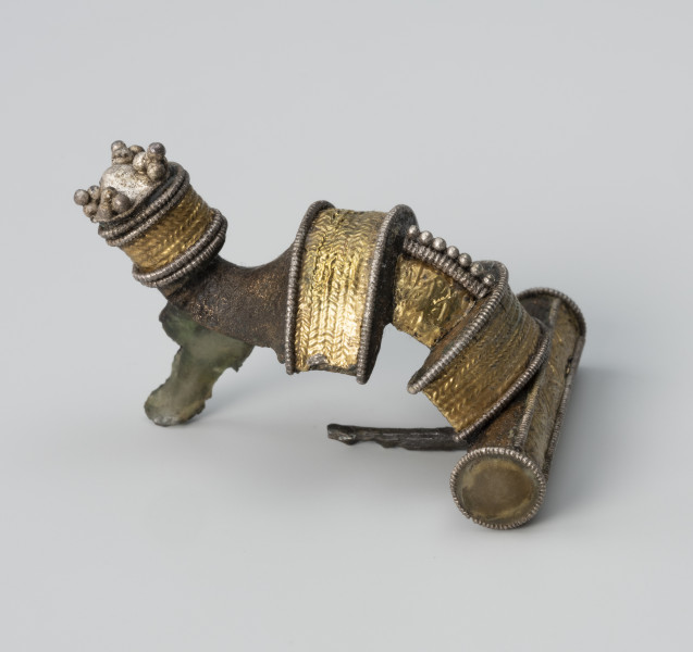 Fibula zdobiona aplikacjami - ujęcie z prawej; Fibula z brązu posiada dwa grzebyki, na kabłąku i nad kapturkiem zakrywającym sprężynkę oraz cylindryczny guz na końcu kabłąka. Jest bogato zdobiona na obu grzebykach i kapturku aplikacjami z repusowanej złotej folii oraz srebrnego filigranu.