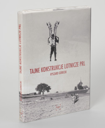 MNS/Sp/1908/4 Ryszard Górecki. Tajne konstrukcje lotnicze PRL - ujęcie z przodu; Obłożony w foliową koszulkę obiekt-książka. Na frontowej stronie okładki czarno-biały fotomontaż z przedstawieniem odwróconego tyłem, unoszącego się w powietrzu żołnierza. Na plecach żołnierza plecak odrzutowy oznakowany szachownicą (symbolem polskich lotniczych sił zbrojnych), na ramionach skrzydła polskiej husarii. Żołnierz unosi się nad zaoranymi polami. Na pierwszym planie po prawej stronie pies, w tle po prawej sylweta kościoła, po lewej zabudowania gospodarcze. Na środku okładki czerwony napis z tytułem publikacji (TAJNE KONSTRUKCJE LOTNICZE PRL) i nazwiskiem autora (RYSZARD GÓRECKI). U dołu okładki czerwone logo wydawnictwa Niewidzialny Znak. Na grzbiecie od góry: logo, tytuł publikacji, nazwisko autora i logo wydawnictwa.