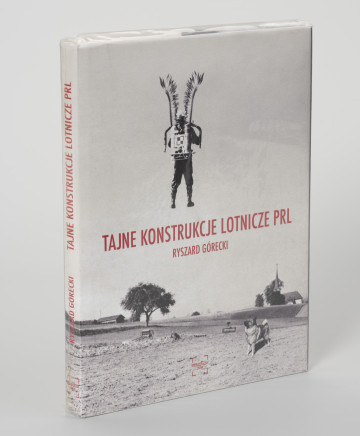 Ryszard Górecki. Tajne konstrukcje lotnicze PRL - ujęcie z przodu; Obłożony w foliową koszulkę obiekt-książka. Na frontowej stronie okładki czarno-biały fotomontaż z przedstawieniem odwróconego tyłem, unoszącego się w powietrzu żołnierza. Na plecach żołnierza plecak odrzutowy oznakowany szachownicą (symbolem polskich lotniczych sił zbrojnych), na ramionach skrzydła polskiej husarii. Żołnierz unosi się nad zaoranymi polami. Na pierwszym planie po prawej stronie pies, w tle po  prawej sylweta kościoła, po lewej zabudowania gospodarcze. Na środku okładki czerwony napis z tytułem publikacji (TAJNE KONSTRUKCJE LOTNICZE PRL) i nazwiskiem autora (RYSZARD GÓRECKI). U dołu okładki czerwone logo wydawnictwa Niewidzialny Znak. Na grzbiecie od góry: logo, tytuł publikacji, nazwisko autora i logo wydawnictwa.