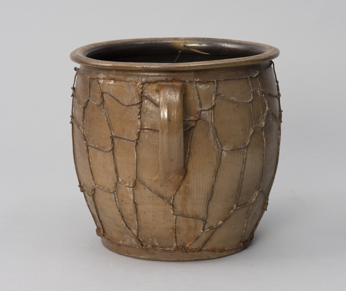 naczynie zasobowe z odrutowaniem - Ujęcie z boku; Duże naczynie zasobowe w kształcie dużego garnka, o słabo wyodrębnionym dnie, szerokim, beczkowatym brzuścu przechodzącym bezpośrednio w kołnierz mocno wygięty na zewnątrz. Brak wyodrębnionej szyjki. Z przodu taśmowe wzdłużne ucho umiejscowione wysoko. Powierzchnia zewnętrzna z wyraźnymi wzdłużnymi rowkami, prawdopodobnie od tokarki. Wewnątrz naczynie powleczone ciemnobrązową emalią. Siatka z drutu oplata całą powierzchnię brzuśca w nieregularny sposób.