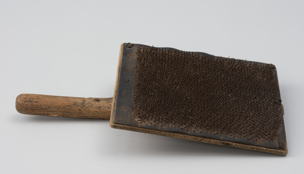 szczotka gręplarska - Ujęcie z boku; Szczotka gręplarska składająca się z części pracującej oraz rączki z lewej. Cześć pracująca wykonana z prostokątnej drewnianej deseczki o naturalnej barwie, do której, z jednej strony przymocowano za pomocą 21 gwoździ skórzaną podkładkę. W podkładkę, od spodu wbite nieregularnie gwoździki o zakrzywionych haczykowato końcach. Na drugiej stronie deseczki, pośrodku, wąska szczelina, w którą wchodzi dość krótka, mocna rączka. Część rączki, która wystaje poza deseczkę, ma przekrój okrągły. Na około ¾ jej wysokości wbity gwóźdź, który zachował się jedynie w części. Na całości widoczne ślady użytkowania.