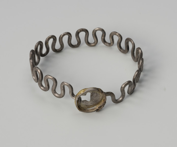 Bransoleta wężykowata - ujęcie z góry; Bransoleta wężykowata ze srebra z owalnym kapsułkowym zapięciem. Elementy zapięcia wycięto ze srebrnej blachy i zlutowano. Obecnie przy zapięciu brakuje przedniej blaszki. Zachowana jest tylna blaszka z T-kształtnym otworem na haczyk. Średnica zamknięcia wynosi 2 cm.