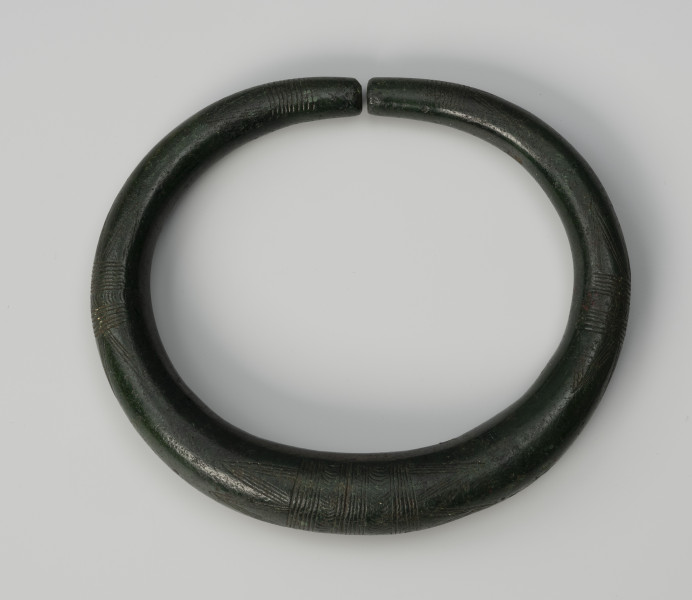 obręcz-nagolennik - Ujęcie z góry; Nagolennik w formie masywnej owalnej, otwartej obręczy o przekroju kolistym, którego średnica zmniejsza się w stronę obydwu prosto ściętych końców. Powierzchnię obręczy zdobi pięć pasm ornamentu złożonego z grup poprzecznych kresek połączonych z kątami wsuwanymi. Całość tworzy motyw tzw. wilczych zębów. Zabytek pokrywa zielona szlachetna patyna.