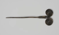 Szpila - Ujęcie z przodu; Szpilę z brązu, należącą do ozdób typu stanomińskiego, charakteryzuje lekko powyginana igła o długości 12,5 cm, która rozszerza się u nasady, tworząc prostokątną płytkę rozdzielającą się na dwa proste ramiona zakończone dwoma czterozwojowymi tarczkami w formie wolut (ślimacznic), na krawędziach których widoczne są szwy odlewnicze. Powierzchnia w dużym stopniu pokryta brunatną patyną.