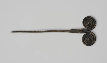 Szpila - Ujęcie z przodu; Szpilę z brązu, należącą do ozdób typu stanomińskiego, charakteryzuje lekko powyginana igła o długości 12,5 cm, która rozszerza się u nasady, tworząc prostokątną płytkę rozdzielającą się na dwa proste ramiona zakończone dwoma czterozwojowymi tarczkami w formie wolut (ślimacznic), na krawędziach których widoczne są szwy odlewnicze. Powierzchnia w dużym stopniu pokryta brunatną patyną.