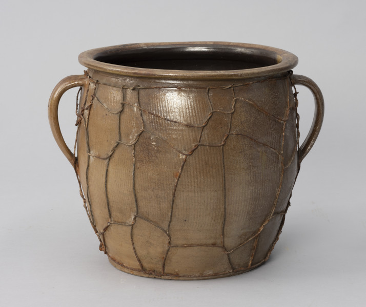 naczynie zasobowe z odrutowaniem - Ujęcie z przodu; Duże naczynie zasobowe w kształcie dużego garnka, o słabo wyodrębnionym dnie, szerokim, beczkowatym brzuścu przechodzącym bezpośrednio w kołnierz mocno wygięty na zewnątrz. Brak wyodrębnionej szyjki. Dwa taśmowe wzdłużne ucha umiejscowione wysoko. Powierzchnia zewnętrzna z wyraźnymi wzdłużnymi rowkami, prawdopodobnie od tokarki. Siatka z drutu oplata całą powierzchnię brzuśca w nieregularny sposób.