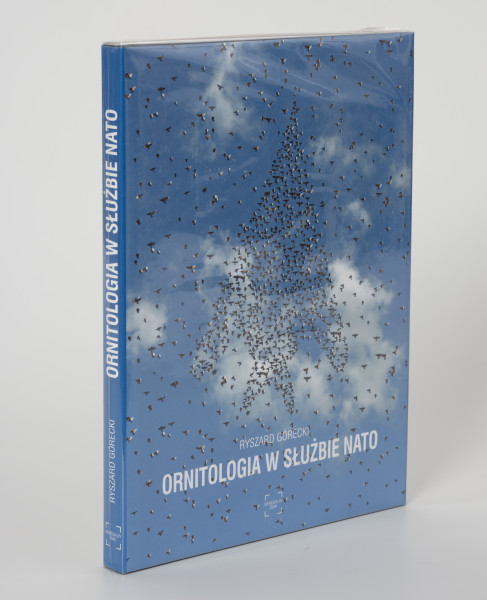 Ryszard Górecki. Ornitologia w służbie NATO - ujęcie z przodu; Obłożony w foliową koszulkę obiekt-książka. Na frontowej stronie niebieskiej okładki fotomontaż z przedstawieniem błękitnego nieba i stada ptaków układającego się w zarys wznoszącego się ku górze bojowego odrzutowca. W tle białe pierzaste chmury. Wokół zarysu samolotu rozproszone na całej powierzchni ptaki. U dołu okładki kolejno białymi literami: nazwisko autora (RYSZARD GÓRECKI), tytuł publikacji (ORNIOTOLOGIA W SŁUŻBIE NATO) i logo wydawnictwa Niewidzialny Znak. Na grzbiecie od góry białymi literami tytuł publikacji, nazwisko autora, logo wydawnictwa. logo, nazwisko autora i tytuł.
