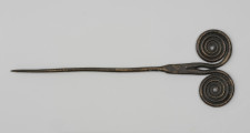Szpila - Ujęcie z tyłu; Szpilę z brązu, należącą do ozdób typu stanomińskiego, charakteryzuje lekko powyginana igła o długości 12,5 cm, która rozszerza się u nasady, tworząc prostokątną płytkę rozdzielającą się na dwa proste ramiona zakończone dwoma czterozwojowymi tarczkami w formie wolut (ślimacznic), na krawędziach których widoczne są szwy odlewnicze. Powierzchnia w dużym stopniu pokryta brunatną patyną.
