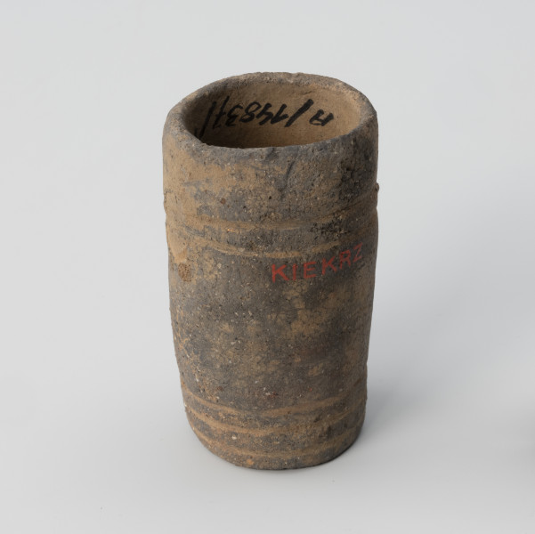 Amfora miniaturowa - ujęcie z boku; Nieprofilowana, miniaturowa amfora bez wyodrębnionego brzuśca, szyjki i wylewu ma kształt walca. Na poziomie 1/3 wysokości naczynka z lewej strony widnieje ślad po uchu. Powierzchnię zdobią dwa dookolne pasma linii rytych: jedno trójliniowe występuje w części przydennej, drugie pasmo dwuliniowe na wysokości uszek. Barwa naczynka jest niejednolita: beżowo-ciemnoszara. Glina zawiera dużą domieszkę miki.