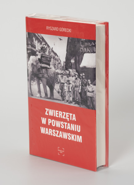 Ryszard Górecki. Zwierzęta w Powstaniu Warszawskim - ujęcie z przodu; Obłożony w foliową koszulkę obiekt-książka. Na frontowej stronie czerwonej okładki od góry: białymi literami nazwisko autora (RYSZARD GÓRECKI), poniżej fotomontaż z przedstawieniem czarno-białej sceny z Powstania Warszawskiego (fragment ulicy przedstawionej w perspektywicznym skrócie, obie pierzeje ciasno zabudowane kilkupiętrowymi kamienicami, na pierwszym planie po prawej grupa uzbrojonych powstańców, po lewej słoń z koszem na grzbiecie, w koszu powstaniec z pistoletem i małpa z granatem, pod zdjęciem białymi literami: tytuł publikacji (ZWIERZĘTA W POWSTANIU WARSZAWSKIM) oraz logo wydawnictwa Niewidzialny Znak.