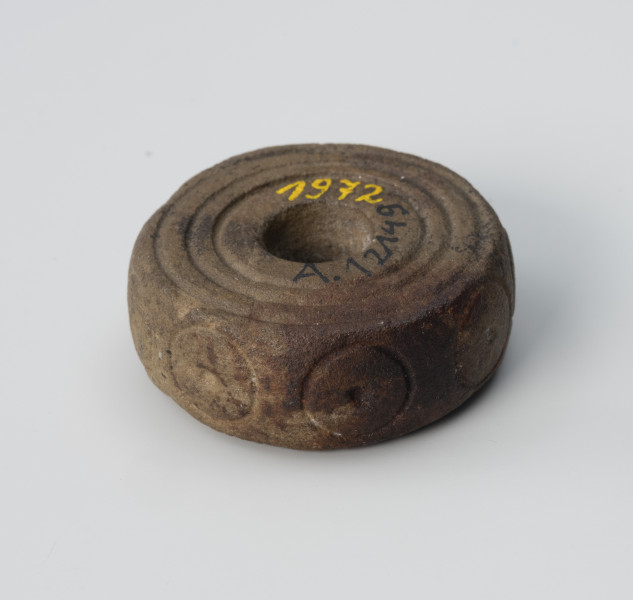 Przęślik - ujęcie z przodu spodu; Krążkowy przęślik z miękkiego surowca skalnego, zdobiony motywem trzech dookolnych linii rytych na biegunach i kółeczkami na największej wydętości.