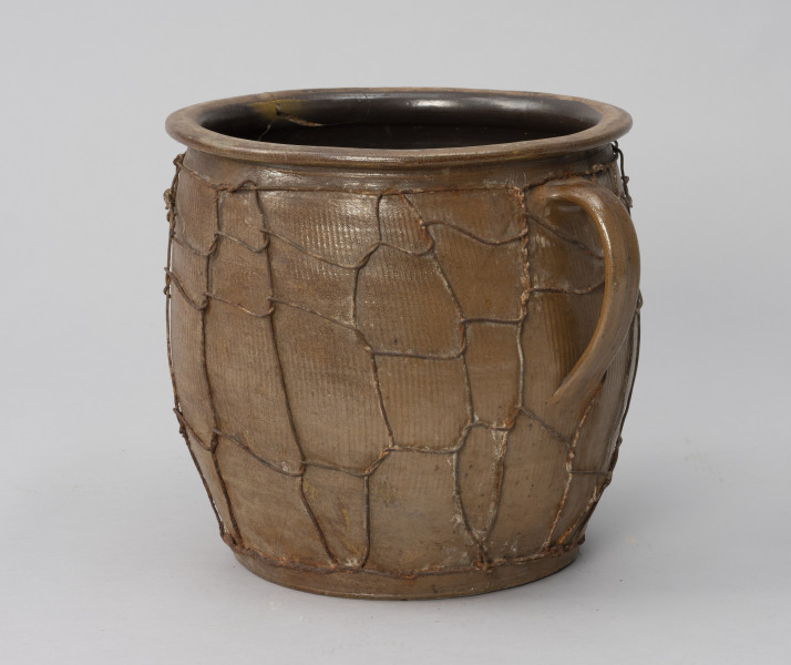 naczynie zasobowe z odrutowaniem - Ujęcie ze skosu z prawej; Duże naczynie zasobowe w kształcie dużego garnka, o słabo wyodrębnionym dnie, szerokim, beczkowatym brzuścu przechodzącym bezpośrednio w kołnierz mocno wygięty na zewnątrz. Brak wyodrębnionej szyjki. Z prawej taśmowe wzdłużne ucho umiejscowione wysoko. Powierzchnia zewnętrzna z wyraźnymi wzdłużnymi rowkami, prawdopodobnie od tokarki. Wewnątrz naczynie powleczone ciemnobrązową emalią. Siatka z drutu oplata całą powierzchnię brzuśca w nieregularny sposób.