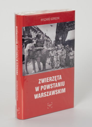 Ryszard Górecki. Zwierzęta w Powstaniu Warszawskim - ujęcie z przodu; Obłożony w foliową koszulkę obiekt-książka. Na frontowej stronie czerwonej okładki od góry: białymi literami nazwisko autora (RYSZARD GÓRECKI), poniżej fotomontaż z przedstawieniem czarno-białej sceny z Powstania Warszawskiego (fragment ulicy przedstawionej w perspektywicznym skrócie, obie pierzeje ciasno zabudowane kilkupiętrowymi kamienicami, na pierwszym planie po prawej grupa uzbrojonych powstańców, po lewej słoń z koszem na grzbiecie, w koszu powstaniec z pistoletem i małpa z granatem, pod zdjęciem białymi literami: tytuł publikacji (ZWIERZĘTA W POWSTANIU WARSZAWSKIM) oraz logo wydawnictwa Niewidzialny Znak. Na grzbiecie od góry: tytuł publikacji, nazwisko autora, logo wydawnictwa.