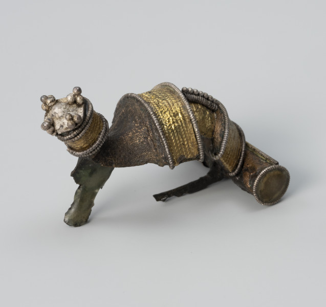 Fibula zdobiona aplikacjami - ujęcie z prawej; Fibula z brązu posiada dwa grzebyki, na kabłąku i nad kapturkiem zakrywającym sprężynkę oraz cylindryczny guz na końcu kabłąka. Jest bogato zdobiona na obu grzebykach i kapturku aplikacjami z repusowanej złotej folii oraz srebrnego filigranu.