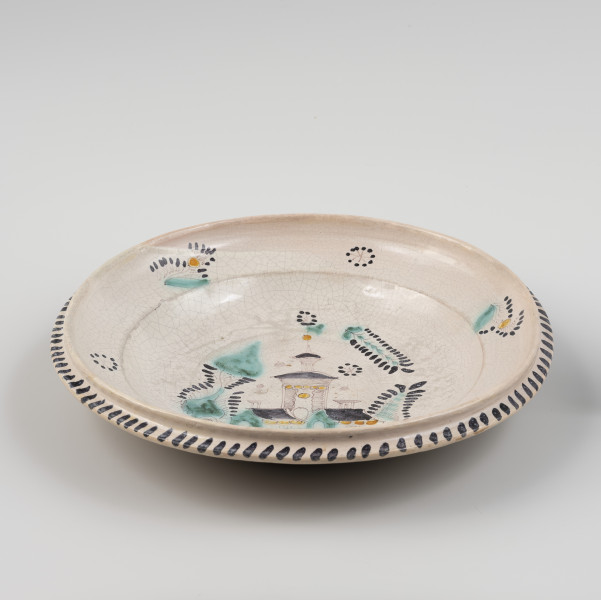 talerz - Ujęcie z przodu; Talerz z czarą naczynia stosunkowo płaską obwiedzioną wyraźnie zaznaczonym wylewem z wysokim rantem i kołnierzem. Szkliwo ołowiowo-cynowe w tonacji szarej pokrywa tylko wewnętrzną stronę i stanowi tło dla odręcznej dekoracji wykonanej tlenkami miedzi, manganu, chromu i antymonu. Trójbarwny, czarno-zielono-żółty malunek przedstawia trzyczęściową budowlę z wieżą. Wokół budowli dekoracja o motywach roślinnych. Wylew zdobiony powtarzającymi się trzykrotnie naprzemiennie wzorami kwiatowymi realistycznymi i schematycznymi. Kołnierz dekorowany skośnymi kreskami. W górnej części wylewu, nad budowlą, wyraźnie cieńsza powłoka szkliwa.