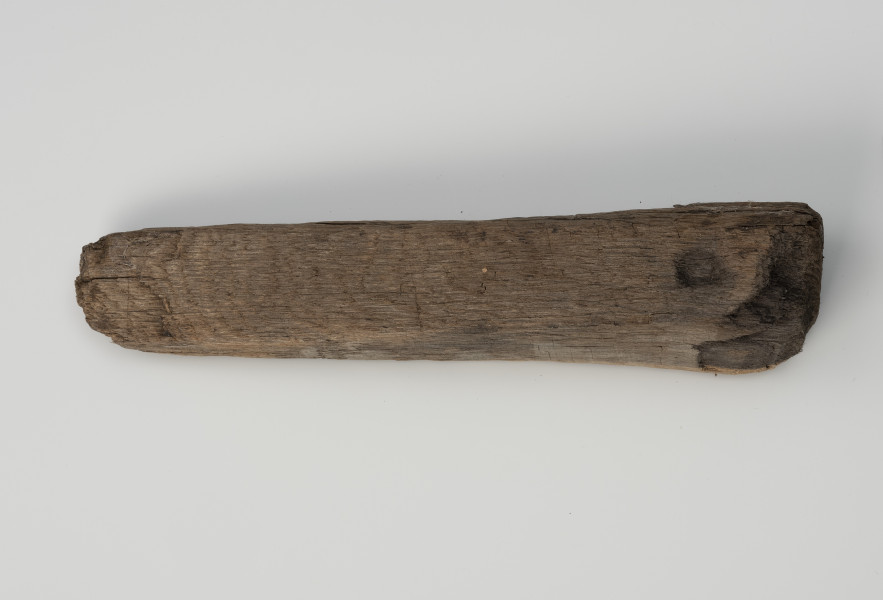 Klin - Ujęcie z góry; Klin w formie graniastosłupa o przekroju zbliżonym do wydłużonego trójkąta równoramiennego. Powierzchnię pozostawiono nieobrobioną, surową i ze śladami cięcia piłą.