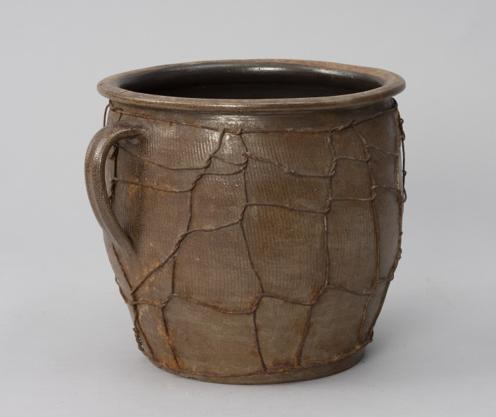 naczynie zasobowe z odrutowaniem - Ujęcie ze skosu z lewej; Duże naczynie zasobowe w kształcie dużego garnka, o słabo wyodrębnionym dnie, szerokim, beczkowatym brzuścu przechodzącym bezpośrednio w kołnierz mocno wygięty na zewnątrz. Brak wyodrębnionej szyjki. Z lewej taśmowe wzdłużne ucho umiejscowione wysoko. Powierzchnia zewnętrzna z wyraźnymi wzdłużnymi rowkami, prawdopodobnie od tokarki. Wewnątrz naczynie powleczone ciemnobrązową emalią. Na brzuścu wzdłużne pęknięcie, które prawdopodobnie było przyczyną odrutowania naczynia. Siatka z drutu oplata całą powierzchnię brzuśca w nieregularny sposób.