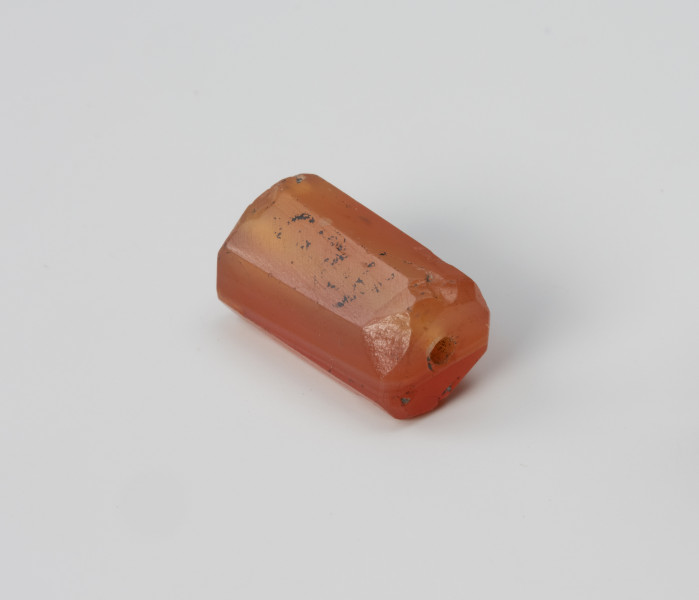 Paciorek wielościenny - ujęcie z przodu; Paciorek z karneolu w kształcie wydłużonego prostopadłościanu, w przekroju ośmioboczny. Kanalik przewiercono wzdłuż dłuższego boku z dwóch stron, z lekką niezbieżnością, a krótsze krawędzie zagładzono.