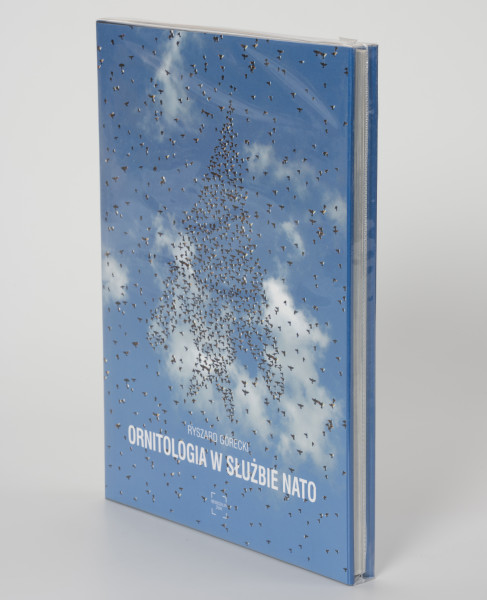 Ryszard Górecki. Ornitologia w służbie NATO - ujęcie z przodu; Obłożony w foliową koszulkę obiekt-książka. Na frontowej stronie niebieskiej okładki fotomontaż z przedstawieniem błękitnego nieba i stada ptaków układającego się w zarys wznoszącego się ku górze bojowego odrzutowca. W tle białe pierzaste chmury. Wokół zarysu samolotu rozproszone na całej powierzchni ptaki. U dołu okładki kolejno białymi literami: nazwisko autora (RYSZARD GÓRECKI), tytuł publikacji (ORNIOTOLOGIA W SŁUŻBIE NATO) i logo wydawnictwa Niewidzialny Znak.