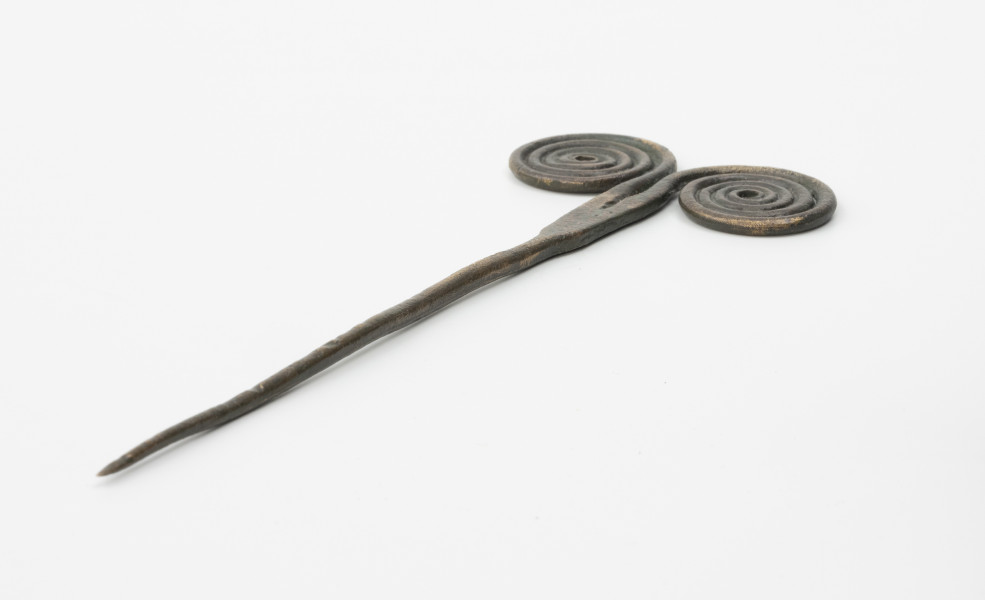 Szpila - Ujęcie ze skosu z drugiej strony; Szpilę z brązu, należącą do ozdób typu stanomińskiego, charakteryzuje lekko powyginana igła o długości 12,5 cm, która rozszerza się u nasady, tworząc prostokątną płytkę rozdzielającą się na dwa proste ramiona zakończone dwoma czterozwojowymi tarczkami w formie wolut (ślimacznic), na krawędziach których widoczne są szwy odlewnicze. Powierzchnia w dużym stopniu pokryta brunatną patyną.