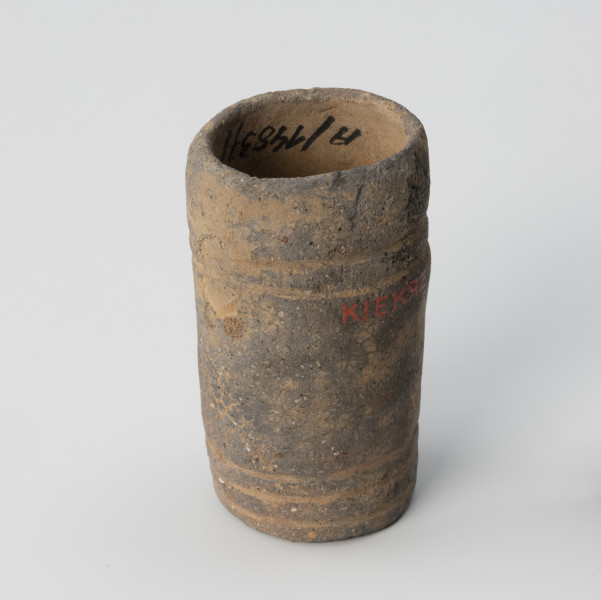 Amfora miniaturowa - ujęcie z boku; Nieprofilowana, miniaturowa amfora bez wyodrębnionego brzuśca, szyjki i wylewu ma kształt walca. Na poziomie 1/3 wysokości naczynka widniej ślad po drugim uchu. Pod uchem znajdują się dwa niewielkie koliste dołki. Powierzchnię zdobią dwa dookolne pasma linii rytych: jedno trójliniowe występuje w części przydennej, drugie pasmo dwuliniowe na wysokości uszek. Barwa naczynka jest niejednolita: beżowo-ciemnoszara. Glina zawiera dużą domieszkę miki.