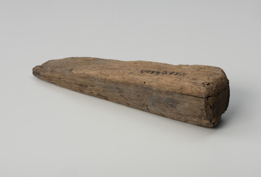 Klin - Ujęcie z tyłu; Klin w formie graniastosłupa o przekroju zbliżonym do wydłużonego trójkąta równoramiennego. Powierzchnię pozostawiono nieobrobioną, surową i ze śladami cięcia piłą.