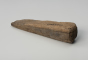 Klin - Ujęcie z tyłu; Klin w formie graniastosłupa o przekroju zbliżonym do wydłużonego trójkąta równoramiennego. Powierzchnię pozostawiono nieobrobioną, surową i ze śladami cięcia piłą.