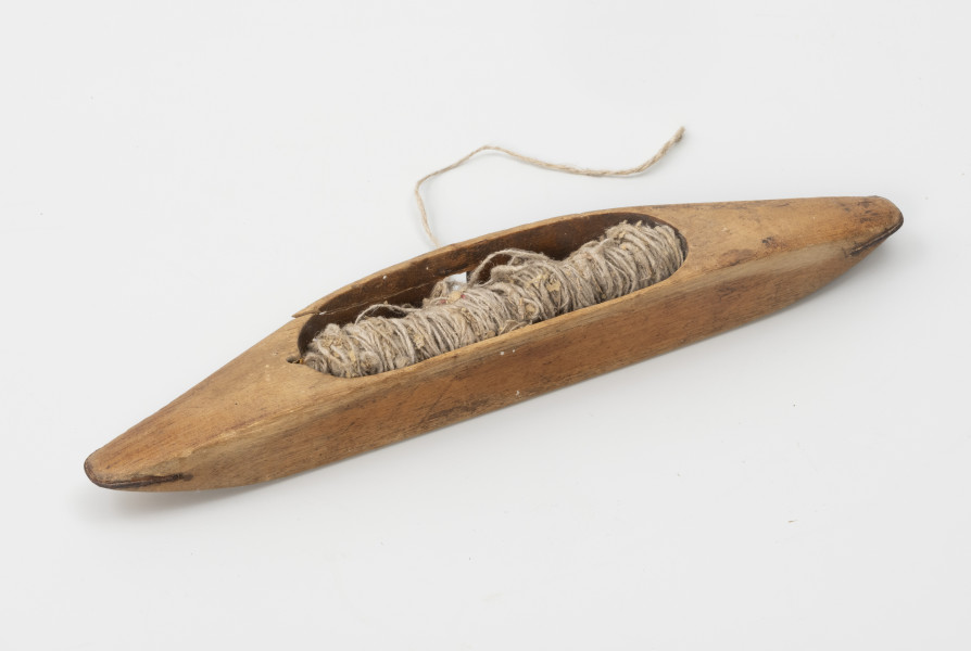 czółenko tkackie - Ujęcie z tyłu ; Czółenko tkackie wykonane z drewna. Korpus kształtem przypomina łódkę. Spód czółenka płaski, ścianki asymetryczne – jedna prosta druga lekko zaokrąglona. Pośrodku czółenka podłużne wgłębienie z zamontowanym metalowym, prostym pręcikiem. Na pręcik nawinięta nić – prawdopodobnie lniana. W ściance tylniej zaokrąglonej, pośrodku kwadratowy, niewielki otwór (oczko), przez który snuta była nić. Pionowe ścianki otworu oraz oba końce czółenka wzmocnione metalem.