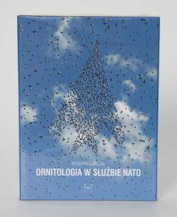 Ryszard Górecki. Ornitologia w służbie NATO - ujęcie z przodu; Obłożony w foliową koszulkę obiekt-książka. Na frontowej stronie niebieskiej okładki fotomontaż z przedstawieniem błękitnego nieba i stada ptaków układającego się w zarys wznoszącego się ku górze bojowego odrzutowca. W tle białe pierzaste chmury. Wokół zarysu samolotu rozproszone na całej powierzchni ptaki. U dołu okładki kolejno białymi literami: nazwisko autora (RYSZARD GÓRECKI), tytuł publikacji (ORNIOTOLOGIA W SŁUŻBIE NATO) i logo wydawnictwa Niewidzialny Znak.
