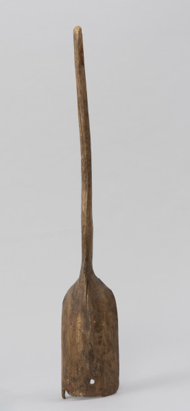 szufla do ziarna - Ujęcie z tyłu; Szufla do ziarna wykonana z jednego kawałka drewna. Część pracująca wąska i długa, Ostrze łyżki uszkodzone mechanicznie, w dwóch miejscach ślady po sękach. Ścianki boczne w górnej partii łagodnie się zwężają i przechodzą w lekko wygięty i zakończony półokrągło trzonek o przekroju owalnym.