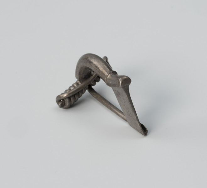 Fibula z wysoką pochewką - ujęcie z tyłu z lewej; Srebrna, niezdobiona zapinka z wysoką pochewką o prostej konstrukcji. Kabłąk ma postać masywnego drutu.