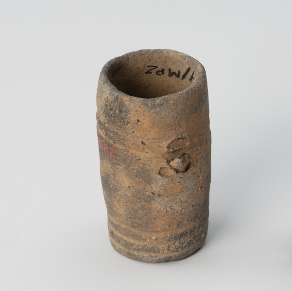 Amfora miniaturowa - ujęcie z tyłu; Nieprofilowana, miniaturowa amfora bez wyodrębnionego brzuśca, szyjki i wylewu ma kształt walca. Na poziomie 1/3 wysokości naczynka widnieją resztki niewielkiego uszka. Drugie uszko nie widoczne z tyłu. Pod każdym z uszek znajdują się dwa niewielkie koliste dołki. Dno jest wklęsłe. Powierzchnię zdobią dwa dookolne pasma linii rytych: jedno trójliniowe występuje w części przydennej, drugie pasmo dwuliniowe na wysokości uszek. Barwa naczynka jest niejednolita: beżowo-ciemnoszara. Glina zawiera dużą domieszkę miki.