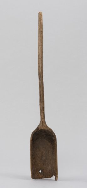 szufla do ziarna - Ujęcie z przodu; Szufla do ziarna wykonana z jednego kawałka drewna. Część pracująca wąska i długa, głęboko wyżłobiona o wyraźnie podniesionych ściankach bocznych. Ostrze łyżki uszkodzone mechanicznie, w dwóch miejscach ślady po sękach. Ścianki boczne w górnej partii łagodnie się zwężają i przechodzą w lekko wygięty i zakończony półokrągło trzonek o przekroju owalnym.