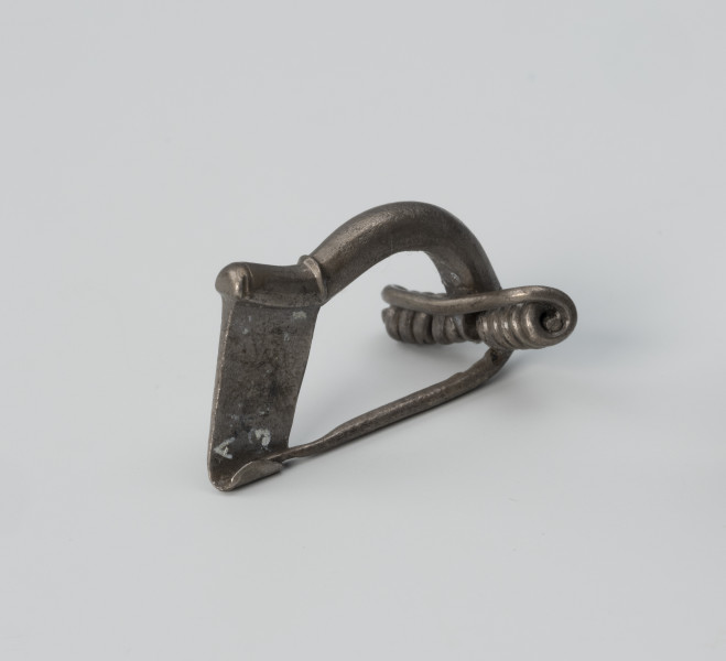 Fibula z wysoką pochewką - ujęcie z tyłu; Srebrna, niezdobiona zapinka z wysoką pochewką o prostej konstrukcji. Kabłąk ma postać masywnego drutu.