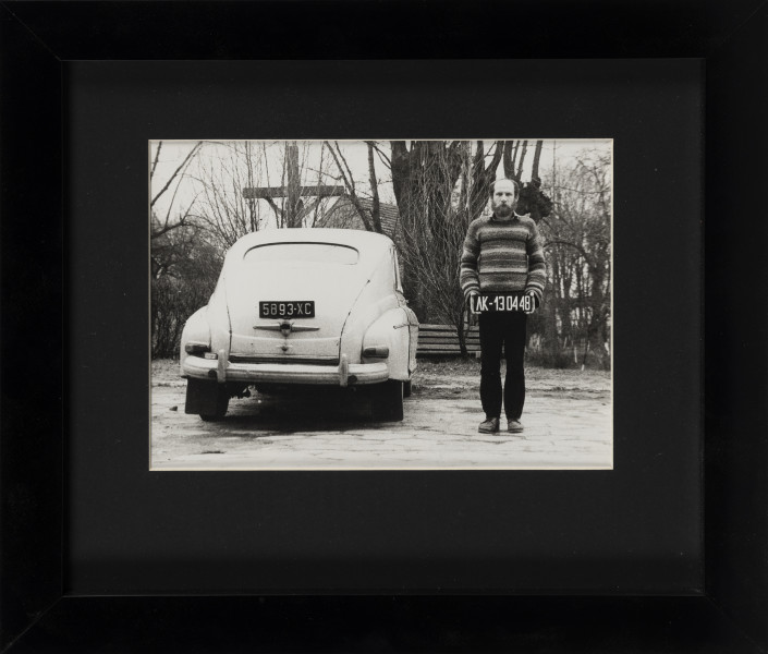 Fotografia; Identyfikacja - ujęcie z przodu; Czarno-biała fotografia w układzie poziomym przedstawiająca scenę zainscenizowaną w plenerze. Na pierwszym planie po prawej stronie sylwetka stojącego mężczyzny  w ujęciu całopostaciowym en face. Mężczyzna trzyma, w opuszczonych wzdłuż ciała rękach, tablicę rejestracyjną z numerem AF-130448. Po lewej tył zaparkowanego białego samochodu z wyeksponowaną tablicą rejestracyjną. W tle drzewa, drewniany krucyfiks i parkowa ławka. Fotografia oprawiona w czarne passe partout oraz czarną, przeszkloną ramę.