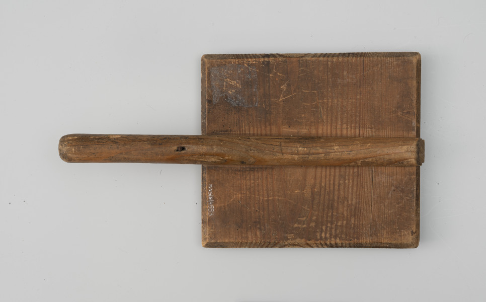 szczotka gręplarska - Ujęcie z tyłu; Szczotka gręplarska. Cześć pracująca wykonana z prostokątnej drewnianej deseczki o naturalnej barwie. Część rączki, która wystaje poza deseczkę, ma przekrój okrągły. Na całości widoczne ślady użytkowania.