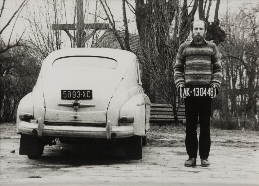 Fotografia; Identyfikacja - ujęcie z przodu; Czarno-biała fotografia w układzie poziomym przedstawiająca scenę zainscenizowaną w plenerze. Na pierwszym planie po prawej stronie sylwetka stojącego mężczyzny  w ujęciu całopostaciowym en face. Mężczyzna trzyma, w opuszczonych wzdłuż ciała rękach, tablicę rejestracyjną z numerem AF-130448. Po lewej tył zaparkowanego białego samochodu z wyeksponowaną tablicą rejestracyjną. W tle drzewa, drewniany krucyfiks i parkowa ławka.