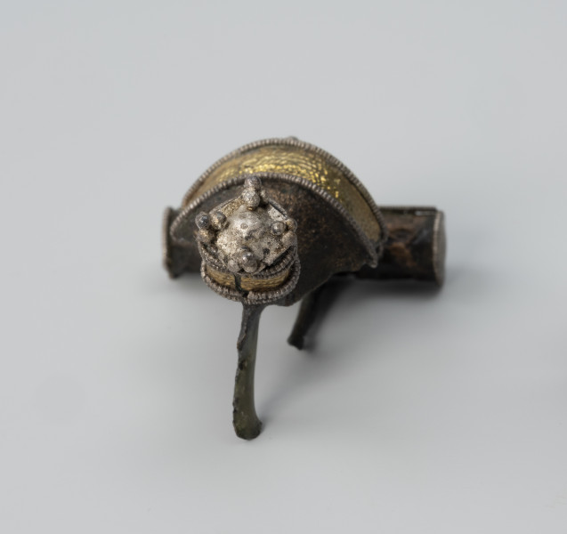 Fibula zdobiona aplikacjami - ujęcie z tyłu; Fibula z brązu. Posiada cylindryczny guz na końcu kabłąka. Jest bogato aplikacjami z repusowanej złotej folii oraz srebrnego filigranu.