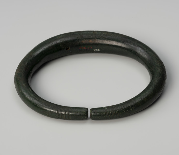 obręcz-nagolennik - Ujęcie z tyłu; Nagolennik w formie masywnej owalnej, otwartej obręczy o przekroju kolistym, którego średnica zmniejsza się w stronę obydwu prosto ściętych końców. Powierzchnię obręczy zdobi pięć pasm ornamentu złożonego z grup poprzecznych kresek połączonych z kątami wsuwanymi. Całość tworzy motyw tzw. wilczych zębów. Zabytek pokrywa zielona szlachetna patyna.