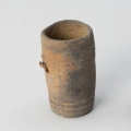 Amfora miniaturowa - ujęcie z drugiego boku; Nieprofilowana, miniaturowa amfora bez wyodrębnionego brzuśca, szyjki i wylewu ma kształt walca. Na poziomie 1/3 wysokości naczynka l lewej strony widnieją resztki niewielkiego uszka. Pod każdym z uszek znajdują się dwa niewielkie koliste dołki. Powierzchnię zdobią dwa dookolne pasma linii rytych: jedno trójliniowe występuje w części przydennej, drugie pasmo dwuliniowe na wysokości uszek. Barwa naczynka jest niejednolita: beżowo-ciemnoszara. Glina zawiera dużą domieszkę miki.