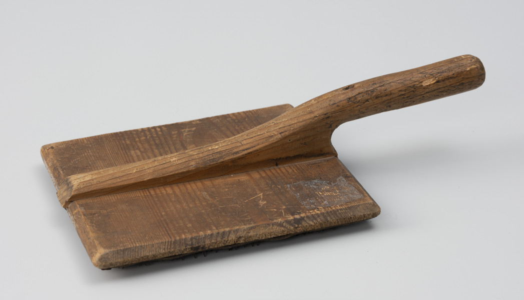szczotka gręplarska - Ujęcie z boku od góry; Szczotka gręplarska składająca się z części pracującej oraz rączki. Cześć pracująca wykonana z prostokątnej drewnianej deseczki o naturalnej barwie. 
Na całości widoczne ślady użytkowania.