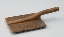 szczotka gręplarska - Ujęcie z boku od góry; Szczotka gręplarska składająca się z części pracującej oraz rączki. Cześć pracująca wykonana z prostokątnej drewnianej deseczki o naturalnej barwie. 
Na całości widoczne ślady użytkowania.