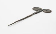 Szpila - Ujęcie ze skosu; Szpilę z brązu, należącą do ozdób typu stanomińskiego, charakteryzuje lekko powyginana igła o długości 12,5 cm, która rozszerza się u nasady, tworząc prostokątną płytkę rozdzielającą się na dwa proste ramiona zakończone dwoma czterozwojowymi tarczkami w formie wolut (ślimacznic), na krawędziach których widoczne są szwy odlewnicze. Powierzchnia w dużym stopniu pokryta brunatną patyną.