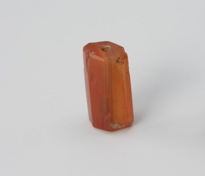 Paciorek wielościenny - ujęcie z przodu; Paciorek z karneolu w kształcie wydłużonego prostopadłościanu, w przekroju ośmioboczny. Kanalik przewiercono wzdłuż dłuższego boku z dwóch stron, z lekką niezbieżnością, a krótsze krawędzie zagładzono.
