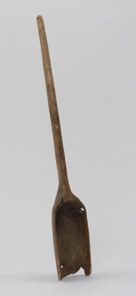 szufla do ziarna - Ujęcie ze skosu z lewej; Szufla do ziarna wykonana z jednego kawałka drewna. Część pracująca wąska i długa, głęboko wyżłobiona o wyraźnie podniesionych ściankach bocznych. Ostrze łyżki uszkodzone mechanicznie, w dwóch miejscach ślady po sękach. Ścianki boczne w górnej partii łagodnie się zwężają i przechodzą w lekko wygięty i zakończony półokrągło trzonek o przekroju owalnym.