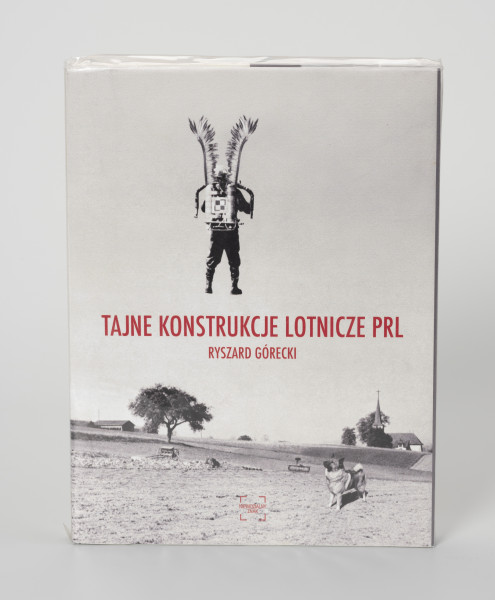 Ryszard Górecki. Tajne konstrukcje lotnicze PRL - ujęcie z przodu; Obłożony w foliową koszulkę obiekt-książka. Na frontowej stronie okładki czarno-biały fotomontaż z przedstawieniem odwróconego tyłem, unoszącego się w powietrzu żołnierza. Na plecach żołnierza plecak odrzutowy oznakowany szachownicą (symbolem polskich lotniczych sił zbrojnych), na ramionach skrzydła polskiej husarii. Żołnierz unosi się nad zaoranymi polami. Na pierwszym planie po prawej stronie pies, w tle po  prawej sylweta kościoła, po lewej zabudowania gospodarcze. Na środku okładki czerwony napis z tytułem publikacji (TAJNE KONSTRUKCJE LOTNICZE PRL) i nazwiskiem autora (RYSZARD GÓRECKI). U dołu okładki czerwone logo wydawnictwa Niewidzialny Znak.