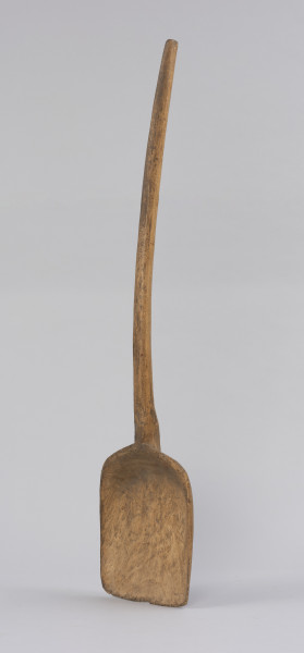 szufla do ziarna - Ujęcie ze skosu od przodu; Szufla do ziarna wykonana z jednego kawałka drewna o szerokiej i płaskiej części pracującej u dołu, której boki i górną krawędź pozostawiono grubsze. Krawędź ta przechodzi łagodnie w łukowato wygięty trzonek, zakończony prosto. Na całej powierzchni narzędzia występują ślady po drewnojadach.