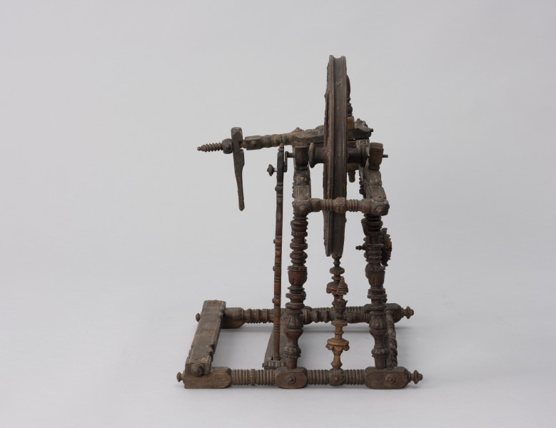 Kołowrotek poziomy - ujęcie z tyłu; Kołowrotek  poziomy o napędzie nożnym. Wykonany z drewna. Elementy kołowrotka łączone na wpusty. Koło napędowe osadzone w podłużnej, poziomej ramie. Wykonana z metalu oś koła oparta na  wspornikach o kształcie. Pierwotnie na drugim końcu ramy mocowany zespół wrzeciona (zachowała się jedynie jego ruchoma podpora). Rama ustawiona na czterech, lekko pochylonych ku środkowi, toczonych na tokarce podpórkach. Pedał w postaci prostej, profilowanej na końcu deseczki połączony z korbą koła napędowego za pomocą drewnianego korbowodu. Całość bogato zdobiona.